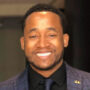 A man with short curly hair and a beard is smiling. He is wearing a dark plaid suit jacket, a black shirt, and a dark tie. The background is softly blurred.