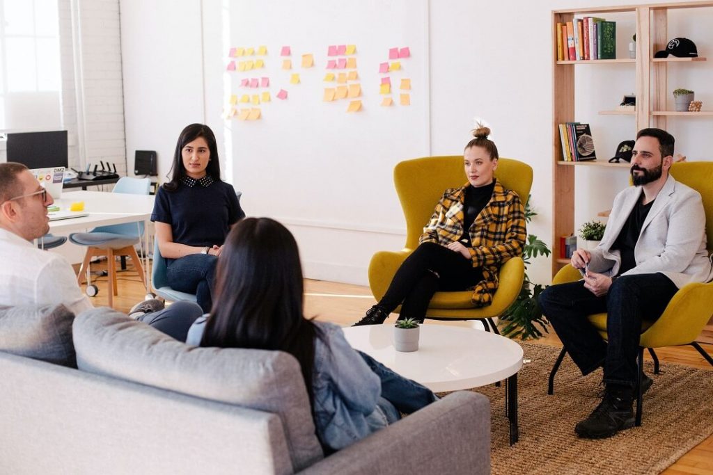 Five people sit in a circle in a modern office space, engaged in conversation. Colorful sticky notes are on the wall behind them, and shelves with books and decor are visible in the background.