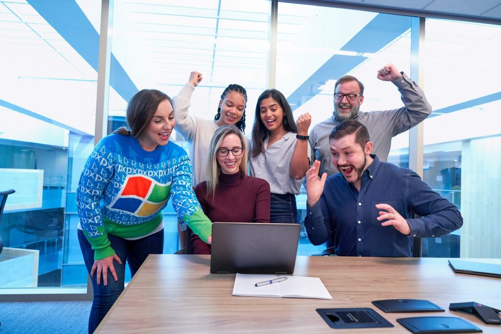 Six colleagues gather around a laptop in a bright office, cheering and smiling excitedly, suggesting they have received good news or achieved a goal together. A notepad and tablet are on the table.