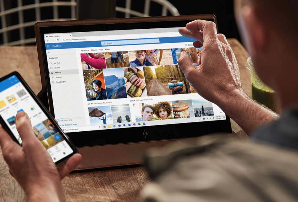 A person uses a smartphone and a touchscreen laptop displaying a photo gallery with multiple images in a web browser, sitting at a wooden table.