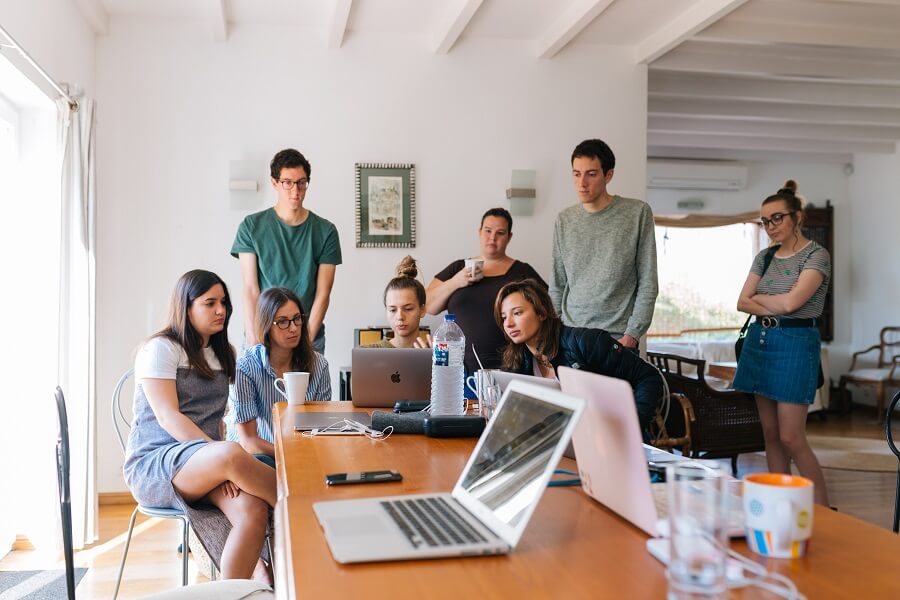 A group of young adults gathers around a table with laptops and drinks, focusing intently on a shared screen in a bright, casual room with large windows and wooden beams.