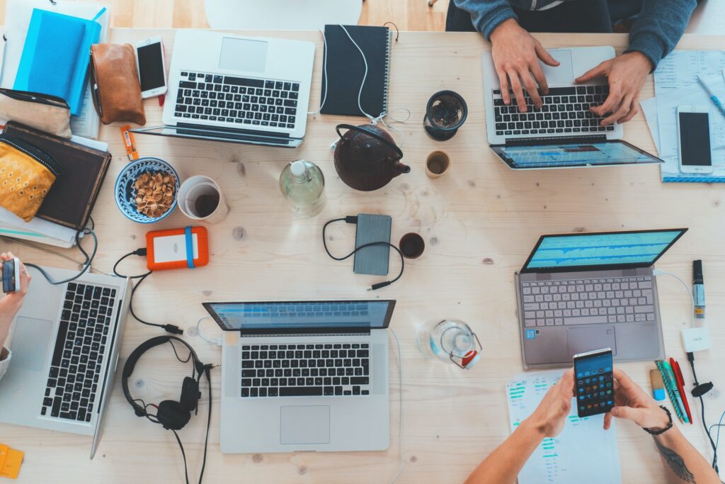 A group of people working at a table with laptops, notebooks, snacks, headphones, water bottles, and a teapot. Hands are typing, writing, and using a smartphone, suggesting a collaborative work or study session.