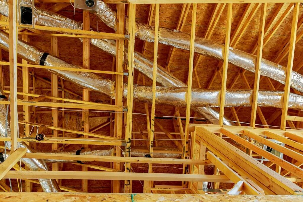 Exposed wooden roof trusses and beams inside a building under construction, with shiny HVAC ductwork running through the ceiling. No walls or ceiling panels have been installed yet.