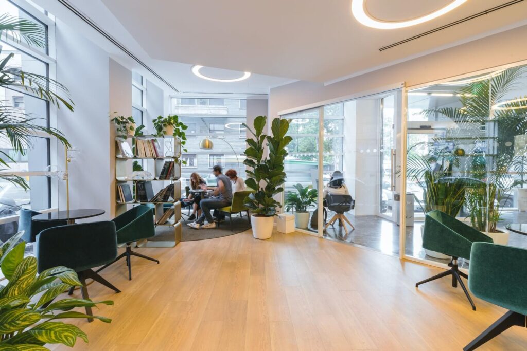 Modern office lounge with green plants, bookshelves, and glass walls. Four people are having a meeting at a round table in the background, while green chairs and natural light fill the space.