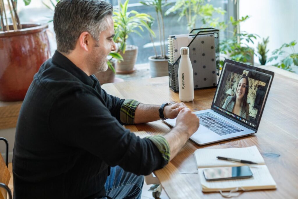 A man sitting at a wooden desk works on a laptop, video chatting with a woman. The desk has a notebook, smartphone, and water bottle. There are plants and sunlight in the background.