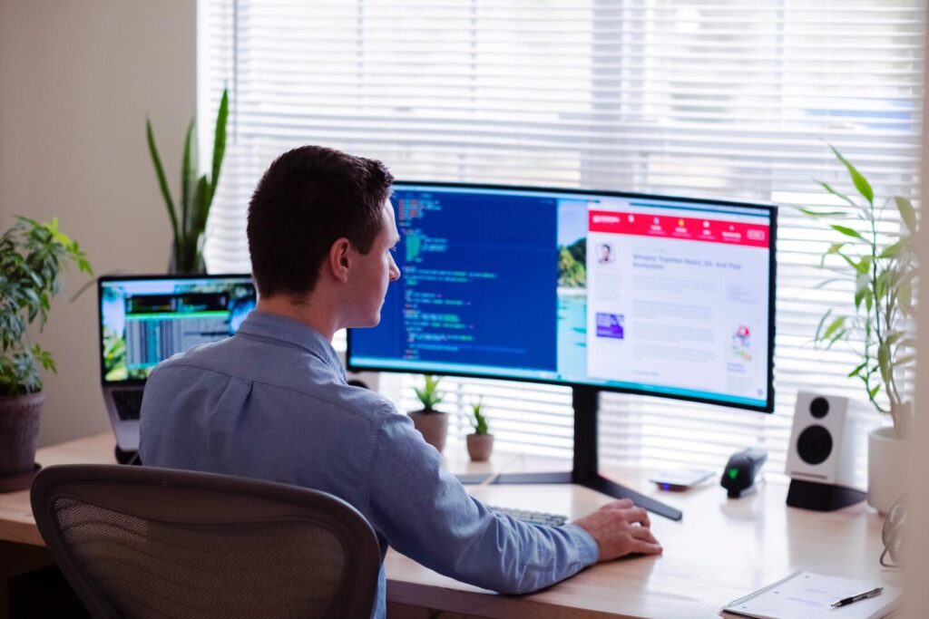 A man sits at a desk working on a computer with dual monitors displaying code and a website. The workspace has potted plants, a laptop, a notebook, and sunlight coming through blinds.