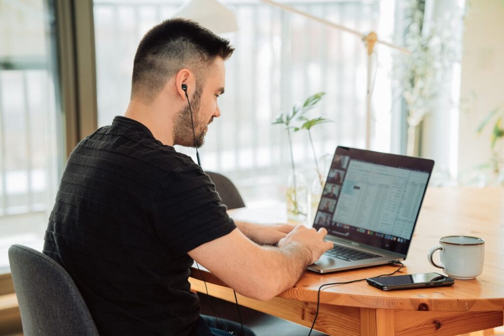 A man with short dark hair, wearing a black shirt and earphones, works on a laptop at a wooden table. A smartphone, coffee mug, and plant are nearby, with windows and natural light in the background.