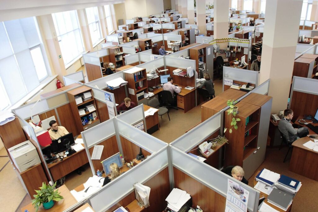 An office with rows of cubicles, each occupied by people working at desks with computers, phones, and paperwork. The workspace has bright lighting and large windows along one wall.