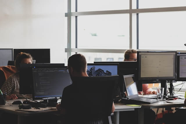 A group of people working at desks with multiple computer monitors in a bright, modern office with large windows.