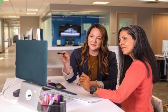 Two women sit at a desk in an office, looking at a computer screen. One woman points at the monitor while the other listens attentively. Office supplies and an open workspace are visible in the background.