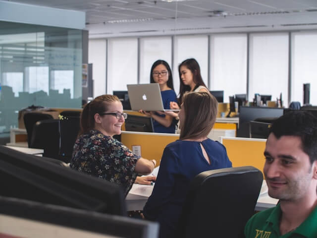 Five people are in a modern office; three women sit and talk at desks while two women stand nearby, one holding a laptop. They appear to be collaborating in a bright, open-plan workspace.