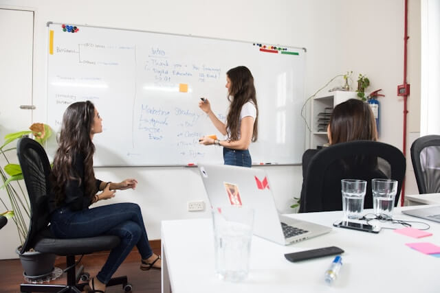 Three women are in an office; one stands and writes on a whiteboard while the others sit and listen. The board displays various notes. Laptops, glasses of water, and office supplies are on the desk in the foreground.